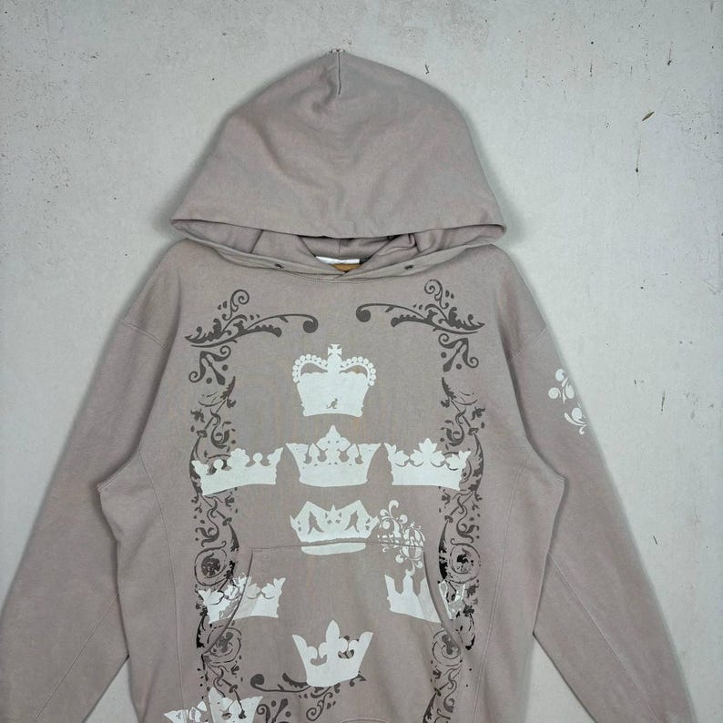 May include: A light taupe hoodie with a drawstring hood and a kangaroo pocket. The front features a design of white crowns and decorative flourishes. The long sleeves have a simple design. The hoodie is made of a soft, comfortable material.