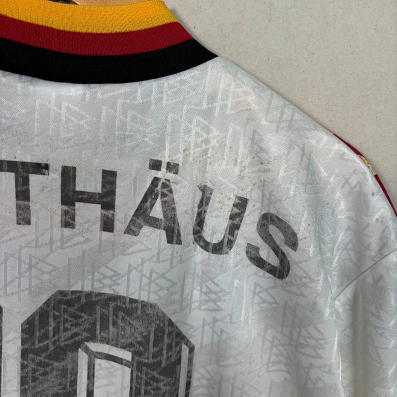 May include: A white football shirt with the name "TH&Auml;US" printed in grey. The shirt features a geometric pattern and a collar with black, red, and yellow stripes. The number "6" is visible.