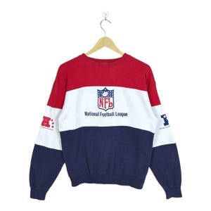 May include: A long-sleeve crewneck sweatshirt featuring a color-blocked design in red, white, and navy blue. The center chest displays the NFL shield logo and the text National Football League. Each sleeve features a small graphic with stars.