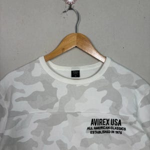 May include: A white and light grey camouflage print long-sleeve shirt with a crew neckline. The chest features black embroidered text that reads AVIREX USA ALL AMERICAN CLASSICS ESTABLISHED IN 1975. The shirt is displayed on a wooden hanger.