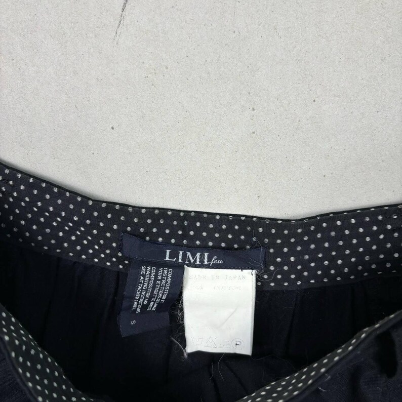 Op de afbeelding: Close-up van een zwarte broek met een witte stippelrand. De broek heeft een label met de tekst "LIMI feu" en bevat wasvoorschriften. De broek is maat S.