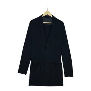 Może przedstawiać: Czarny blazer o wydłużonym kroju, z klapami z wycięciami, zapięciem na trzy guziki i dwiema przednimi kieszeniami. Blazer wisi na drewnianym wieszaku. Rękawy są długie.