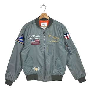 May include: A gray bomber jacket with multiple patches, including the text "FLYING TIGERS" and "P-40 Warhawk". The jacket has a zipper closure, ribbed cuffs, and a collar. The interior lining is orange.
