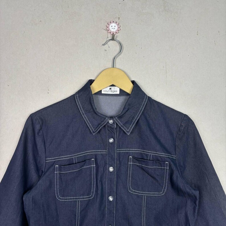 May include: Dark blue denim shirt with white stitching details. Features a collared neckline, two front pockets, and button closures. The shirt is hanging on a wooden hanger.