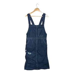 Può includere: Un abito con bretelle in denim blu scuro con spalline regolabili, una tasca sul petto e tasche anteriori. L'abito ha un orlo al ginocchio e un piccolo disegno a spruzzi di vernice bianca e blu sul lato inferiore sinistro. È appeso a una gruccia di legno.