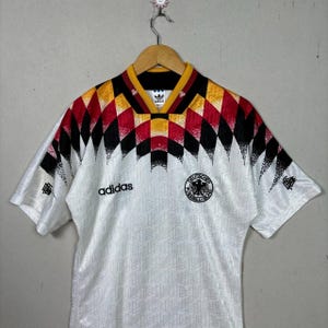 May include: White football shirt with a geometric pattern in black, red, and yellow on the shoulders and collar. Features the Adidas logo and a German national emblem. Short-sleeved with a textured design.