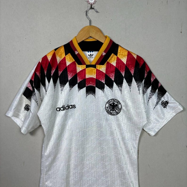 May include: White football shirt with a geometric pattern in black, red, and yellow on the shoulders and collar. Features the Adidas logo and a German national emblem. Short-sleeved with a textured design.