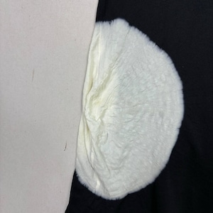 May include: A close-up of a white, fluffy, semi-circular object against a black background. The object has a textured surface and appears to be made of a soft material. To the left, a light beige surface is visible.
