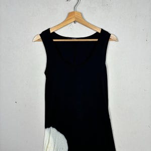 May include: Sleeveless black top with a scoop neckline, hanging on a wooden hanger. The bottom left side of the top features a white, textured design. The top is made of a soft material.