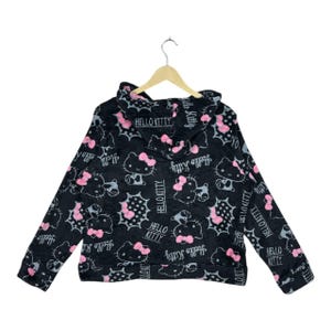 Puede incluir: Sudadera con capucha negra con un patr&oacute;n repetido de personajes de Hello Kitty con lazos rosas y el texto "Hello Kitty". El dise&ntilde;o incluye bocadillos y las palabras "Hello Kitty" en una fuente estilizada.