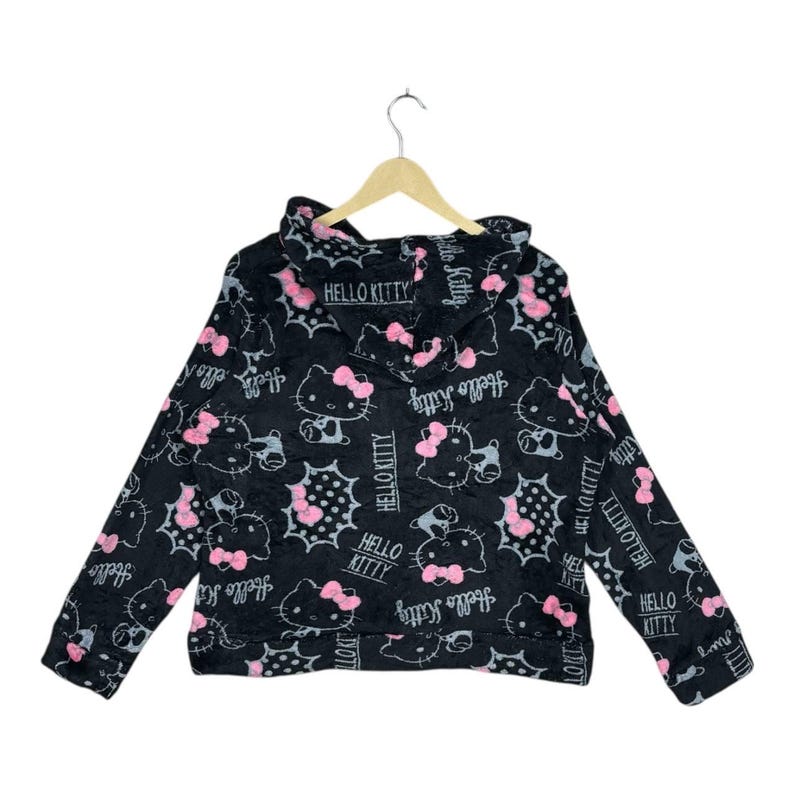 Puede incluir: Sudadera con capucha negra con un patr&oacute;n repetido de personajes de Hello Kitty con lazos rosas y el texto "Hello Kitty". El dise&ntilde;o incluye bocadillos y las palabras "Hello Kitty" en una fuente estilizada.