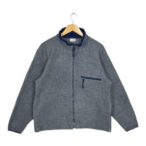 May include: A grey fleece jacket with a full zip, a navy blue collar, and a navy blue pocket on the chest. The jacket is hanging on a wooden hanger. The jacket has a soft, textured appearance.