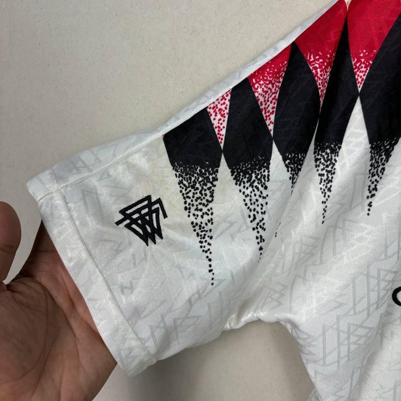 May include: White short-sleeved shirt with a geometric pattern and black and red diamond shapes on the sleeve. The shirt has a small black logo on the sleeve. The fabric has a subtle textured design.