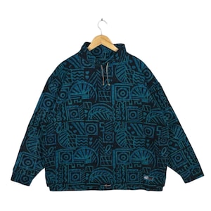 Könnte beinhalten: Ein teal-farbener und schwarzer Fleece-Pullover mit geometrischem Muster. Das langärmlige Oberteil hat einen hohen Kragen mit Teilreißverschluss und ein kleines Logo unten rechts. Das Muster umfasst Kreise, Quadrate und stilisierte Blätter.