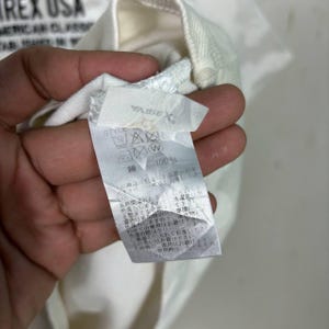 May include: A white garment featuring a fabric care label with the text "AVIREX" and "100%" cotton in Japanese characters. The label includes various laundry instruction symbols, including a washing machine icon, a triangle, and a circle with a W.