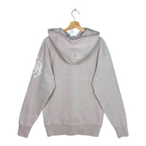 May include: A light taupe hoodie with a drawstring hood and long sleeves. The back of the hoodie is plain, while the left sleeve features a white floral design. The hoodie is hanging on a wooden hanger.