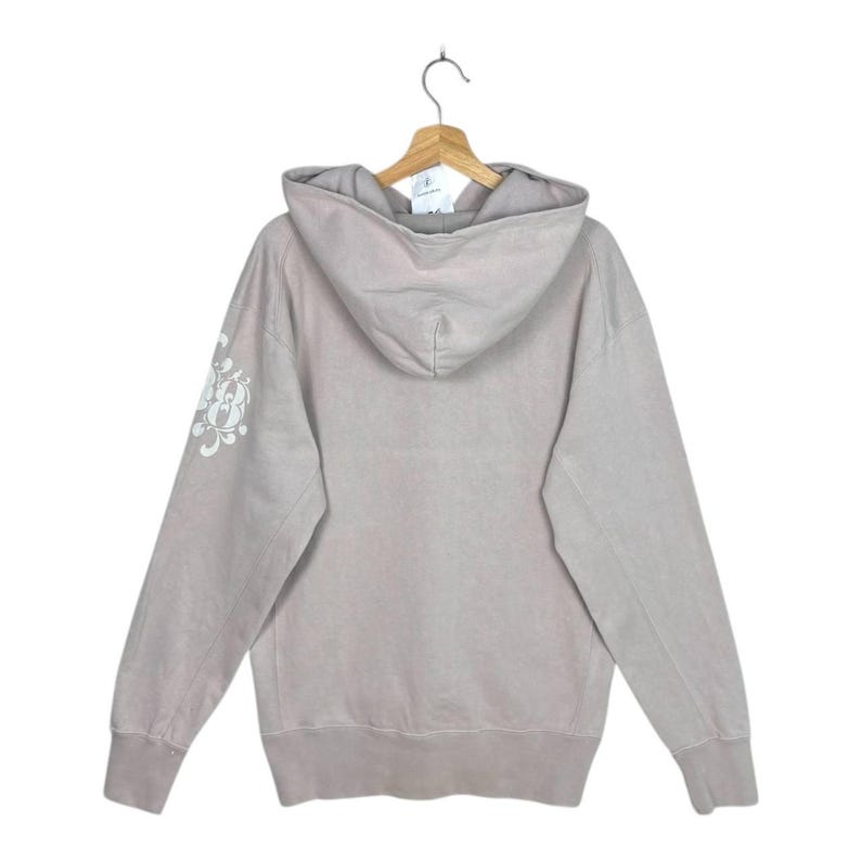 May include: A light taupe hoodie with a drawstring hood and long sleeves. The back of the hoodie is plain, while the left sleeve features a white floral design. The hoodie is hanging on a wooden hanger.