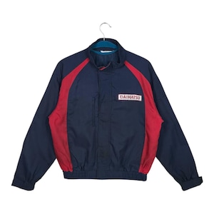 May include: A navy blue and red jacket featuring the word "DAIHATSU" in a white rectangle. The jacket has a stand-up collar, a zip closure, and red panels on the shoulders and sides. A classic design.