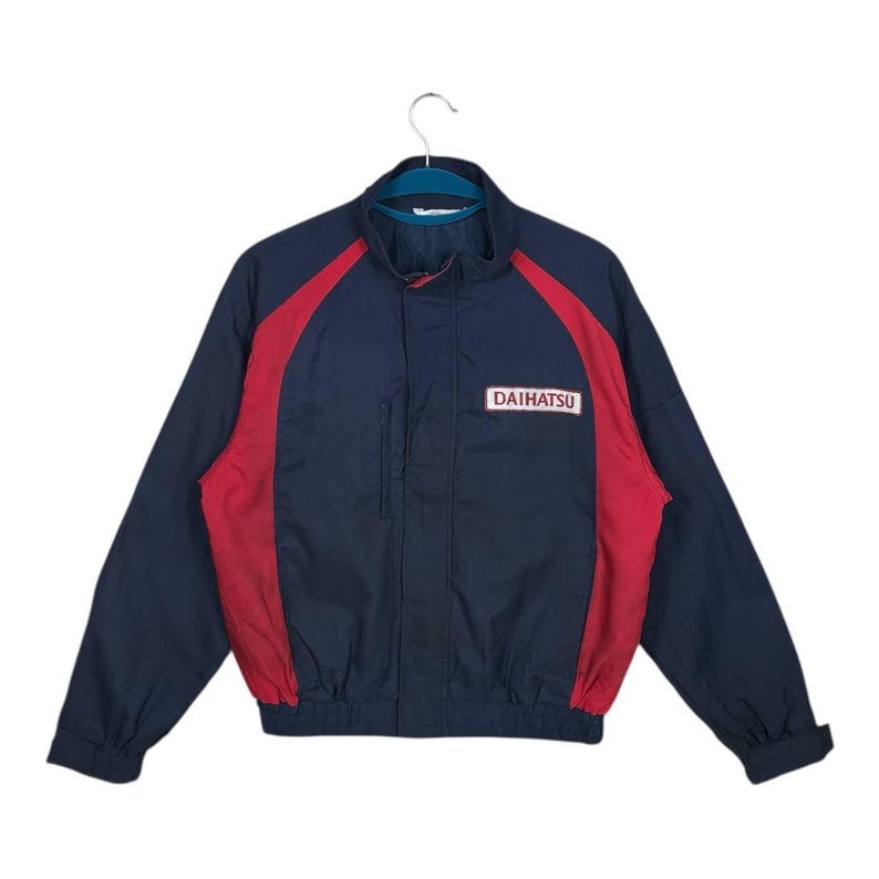May include: Navy blue and red jacket with the word "DAIHATSU" in a white rectangle on the chest. The jacket has a stand-up collar, a zipper closure, and red accents on the shoulders and sides.