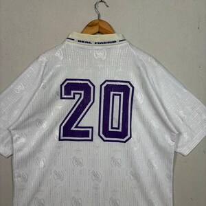 K&ouml;nnte beinhalten: Wei&szlig;es Real Madrid Fu&szlig;balltrikot mit der lila Nummer 20 auf dem R&uuml;cken. Das Trikot hat einen Kragen mit dem blauen Text "REAL MADRID". Der Stoff hat ein dezentes Muster.
