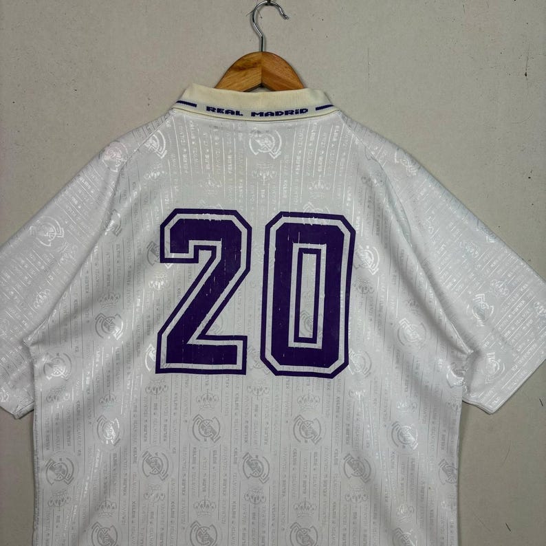 K&ouml;nnte beinhalten: Wei&szlig;es Real Madrid Fu&szlig;balltrikot mit der lila Nummer 20 auf dem R&uuml;cken. Das Trikot hat einen Kragen mit dem blauen Text "REAL MADRID". Der Stoff hat ein dezentes Muster.