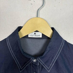 May include: Close-up of a dark blue collared shirt with white stitching detail. The shirt is hanging on a wooden hanger. The label reads "courr&egrave;ges".