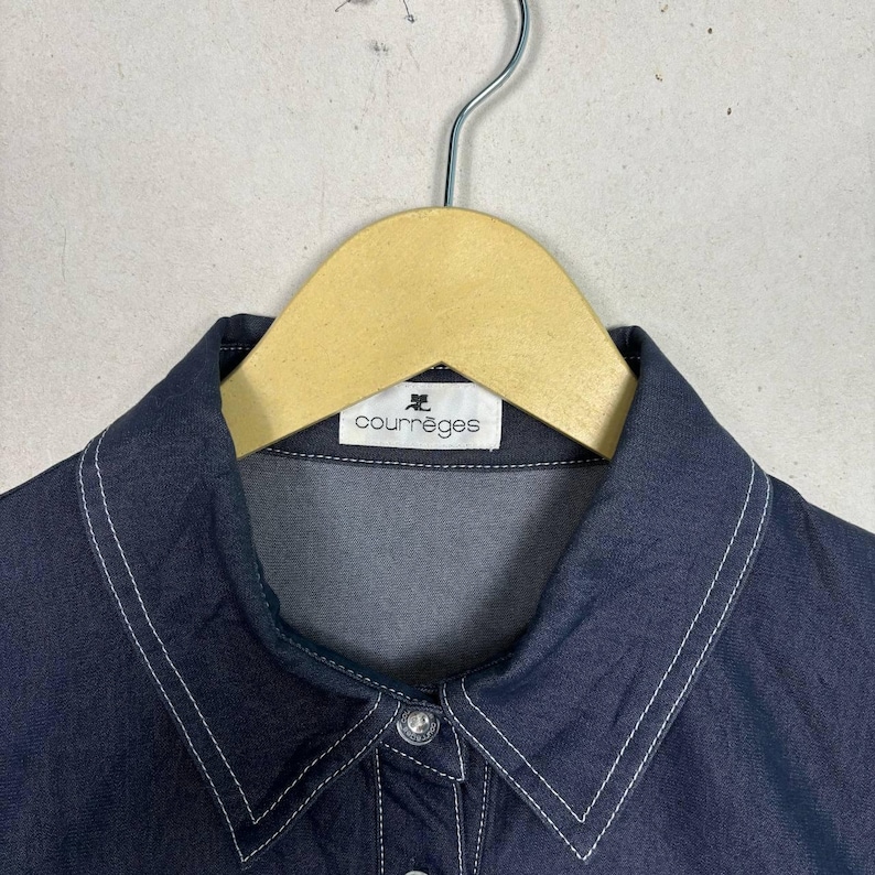 May include: Close-up of a dark blue collared shirt with white stitching detail. The shirt is hanging on a wooden hanger. The label reads "courr&egrave;ges".