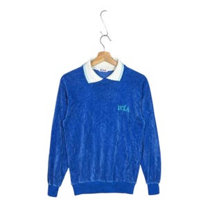 May include: A long-sleeved blue velour pullover shirt featuring a white polo-style collar with light blue stripes. The text UCLA is embroidered in light blue on the left chest. A small white tag with the text UCLA is visible inside the collar.