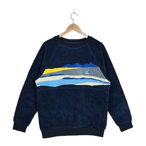 May include: A dark navy blue long-sleeve fleece sweatshirt featuring a textured landscape graphic across the chest. The design includes horizontal bands of yellow, gray, light blue, and cream, with a small square logo patch reading HH.