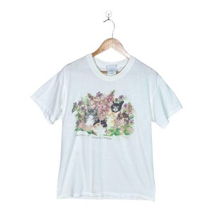 Könnte beinhalten: Ein weißes kurzärmeliges T-Shirt mit einem Grafikdruck von fünf Katzen, umgeben von violetten und grünen Blumen sowie einem Schmetterling. Der Text unter den Katzen lautet "Victoria Canada". Das T-Shirt hängt an einem Holzkleiderbügel.