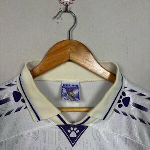 K&ouml;nnte beinhalten: Ein Vintage-Fu&szlig;balltrikot von Real Madrid, wei&szlig; mit lila Akzenten. Das Trikot hat einen cremefarbenen Kragen und Besatz, ein sich wiederholendes Muster und die Logos von Kelme und Real Madrid. Es h&auml;ngt an einem Holzkleiderb&uuml;gel.