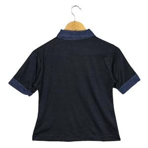May include: Black short-sleeved shirt with a denim collar and denim cuffs. The shirt is hanging on a wooden hanger. The shirt's collar and cuffs are a darker shade of blue than the shirt's body.
