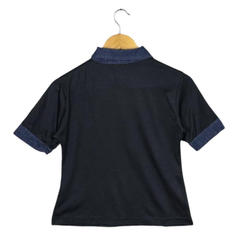 May include: Black short-sleeved shirt with a denim collar and denim cuffs. The shirt is hanging on a wooden hanger. The shirt's collar and cuffs are a darker shade of blue than the shirt's body.