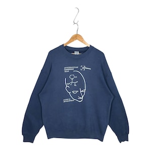 Puede incluir: Sudadera azul marino con texto blanco y un diseño estilizado de cabeza humana. El texto incluye "LEVETIRACETAM SUBMISSION TEAM" y "CLINICAL DEVELOPMENT". La sudadera está colgada de una percha de madera.
