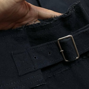 May include: Close-up of a dark navy blue fabric with a silver metal buckle and belt loop. The fabric has a textured appearance with visible stitching and a raw, unfinished edge. The belt is secured with a rectangular buckle.