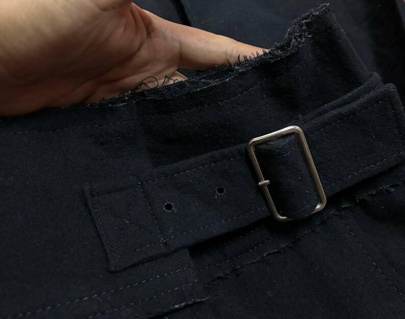May include: Close-up of a dark navy blue fabric with a silver metal buckle and belt loop. The fabric has a textured appearance with visible stitching and a raw, unfinished edge. The belt is secured with a rectangular buckle.