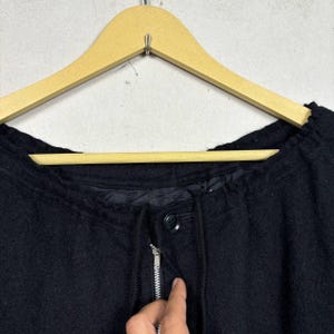 Op de afbeelding: Een zwarte jas met ritssluiting en knoop. De jas hangt aan een houten kleerhanger. De jas heeft een trekkoord rond de halslijn.