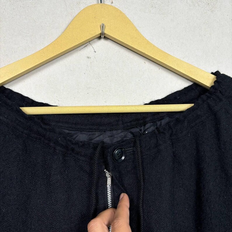 Op de afbeelding: Een zwarte jas met ritssluiting en knoop. De jas hangt aan een houten kleerhanger. De jas heeft een trekkoord rond de halslijn.