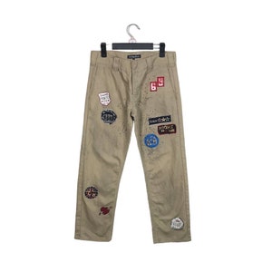 May include: Beige cargo pants with multiple patches and embellishments. The patches feature various designs, including stars, words, and graphics. The pants have a relaxed fit and a straight leg.