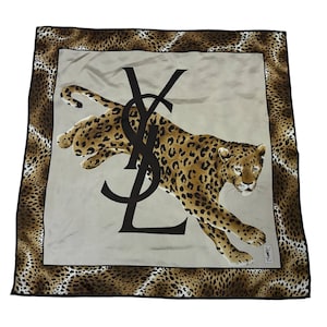 Peut inclure: Un foulard carré en soie présentant un motif central de léopard bondissant sur fond gris clair, surmonté d'un grand logo YSL noir. Le foulard est encadré par une large bordure à motif léopard marron et noir avec des bords noirs.