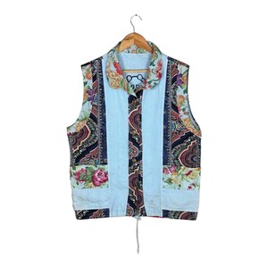 Pode incluir: Um colete sem mangas com um design de patchwork. O colete apresenta um painel central azul claro com listras verticais, ladeado por painéis com estampas florais e paisley. A gola tem um padrão floral e o colete tem um cordão na parte inferior.