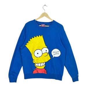 Könnte beinhalten: Blaues Langarm-Sweatshirt mit Bart Simpson mit gelben Haaren, rotem Hemd und einer Sprechblase mit der Aufschrift "I'll be good sometimes". Das Sweatshirt hängt an einem Holzbügel.