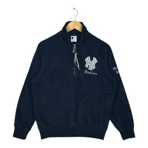 May include: Navy blue Yankees zip-up sweatshirt with the team logo on the chest. The sweatshirt has a high collar and a half-zip closure. The team name "Yankees" is embroidered below the logo. The sleeves are long and the material appears to be a soft fleece.