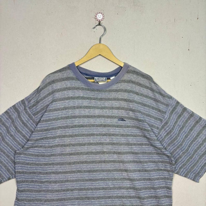 May include: A short-sleeved, striped t-shirt in shades of gray and blue. The shirt features a crew neck and a small logo on the chest. The fabric has a textured appearance, and the stripes run horizontally across the garment.