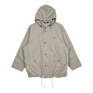 Op de afbeelding: Een beige jas met capuchon, een trekkoord in de zoom en drukknoopsluitingen. De jas heeft twee voorzakken en een klein marineblauw logo op de borst. De jas is gemaakt van een lichtgewicht materiaal.