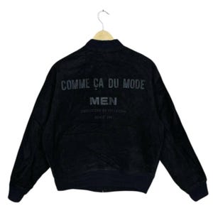 May include: Black bomber jacket with the text "COMME CA DU MODE MEN" on the back. The jacket is suspended from a wooden hanger. The text "PROJECTED BY FIVE FOX" and "SINCE 1981" are also visible.