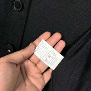 Może przedstawiać: Zbliżenie czarnego ubrania zapinanego na guziki z metką trzymaną w dłoni. Na metce widnieje tekst, w tym "HE-T03-965", procentowa zawartość materiał&oacute;w i instrukcje pielęgnacji. Tkanina ubrania wydaje się być w ciemnym, jednolitym kolorze.