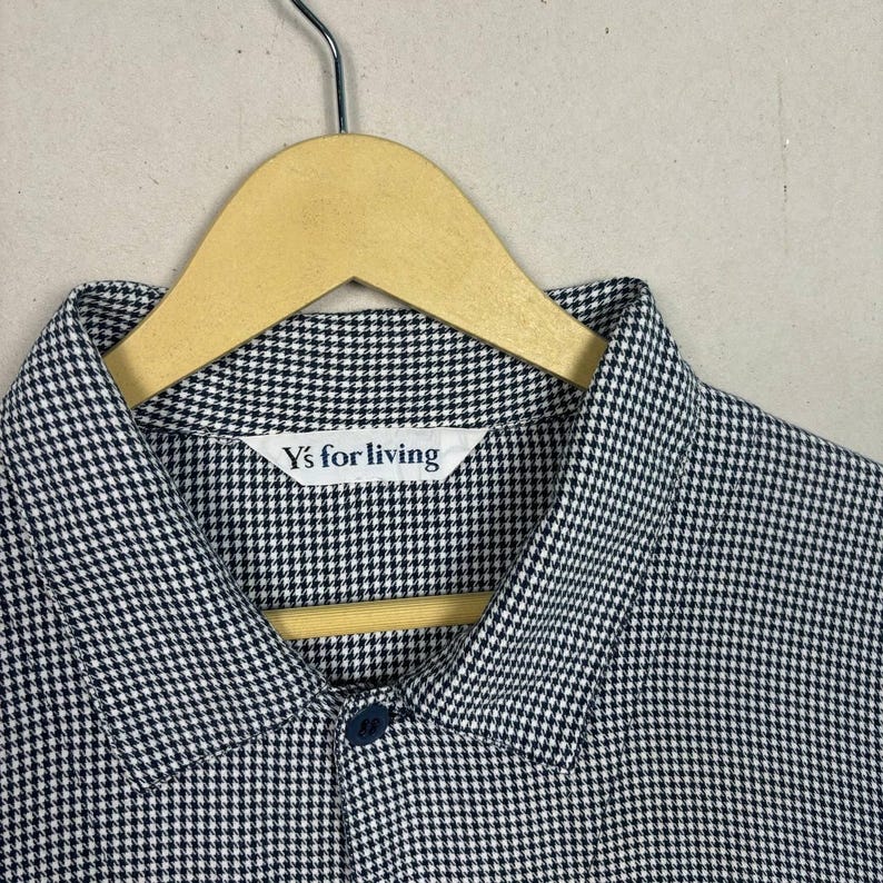 May include: A black and white checkered shirt with a button-down collar, hanging on a wooden hanger. The shirt has a small label that reads "Y's for living". The fabric has a small, repeating pattern.
