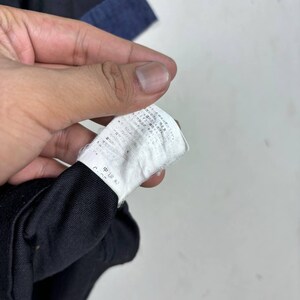 May include: Close-up of a white clothing tag with Japanese text, held by a hand. The tag is attached to a black garment, with a blue fabric visible in the background. The tag appears to be made of a thin, slightly textured material.