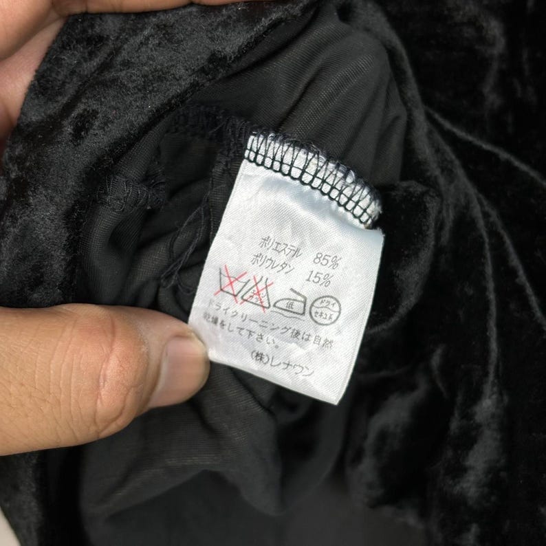 Op de afbeelding: Een zwart stoffen label met witte tekst die de stofsamenstelling beschrijft: 85% polyester en 15% polyurethaan. Het label bevat ook wasinstructies in het Japans.